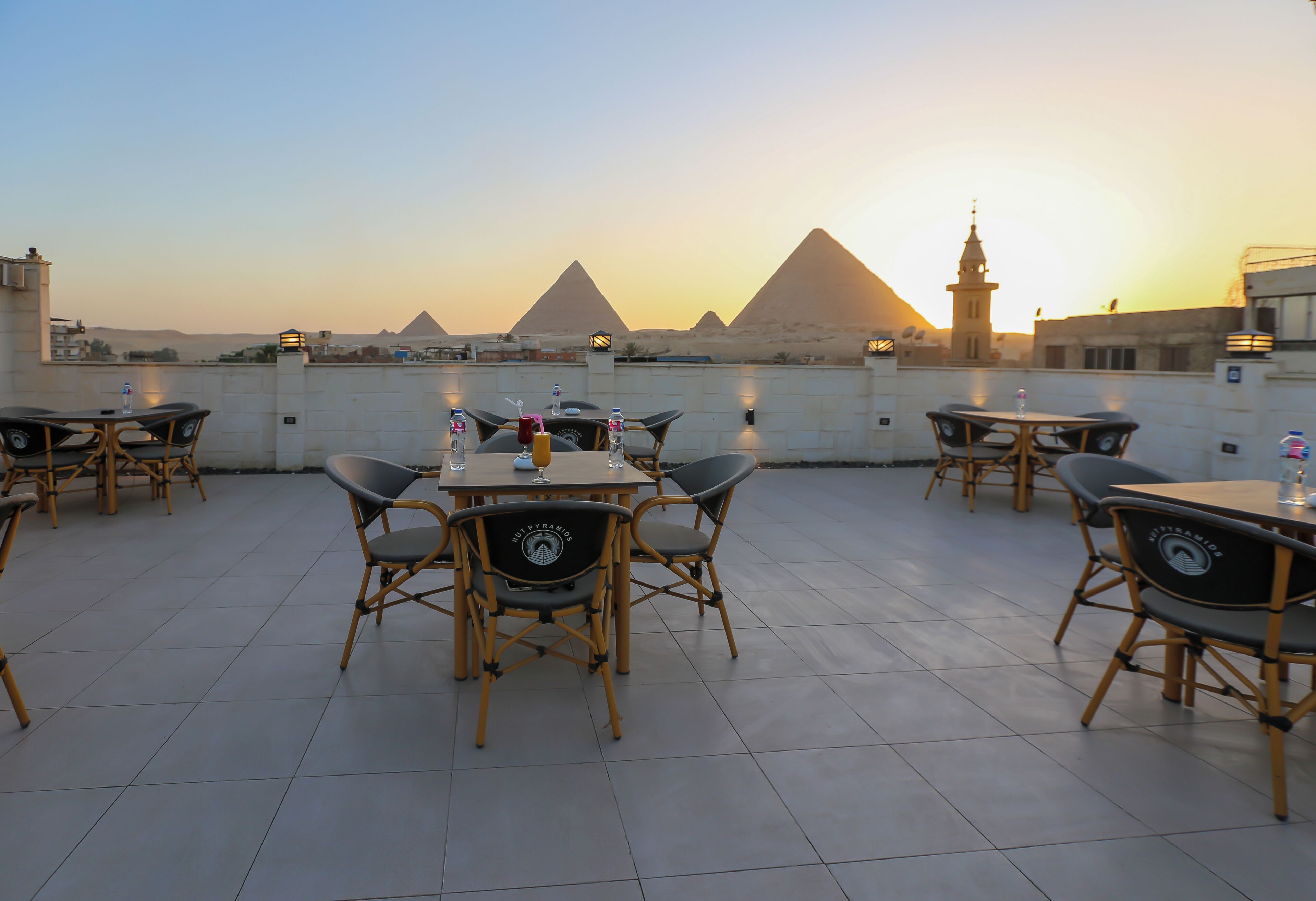 Terraza en la azotea con mesas y sillas, ofreciendo vistas a las pirámides y un minarete al atardecer.
