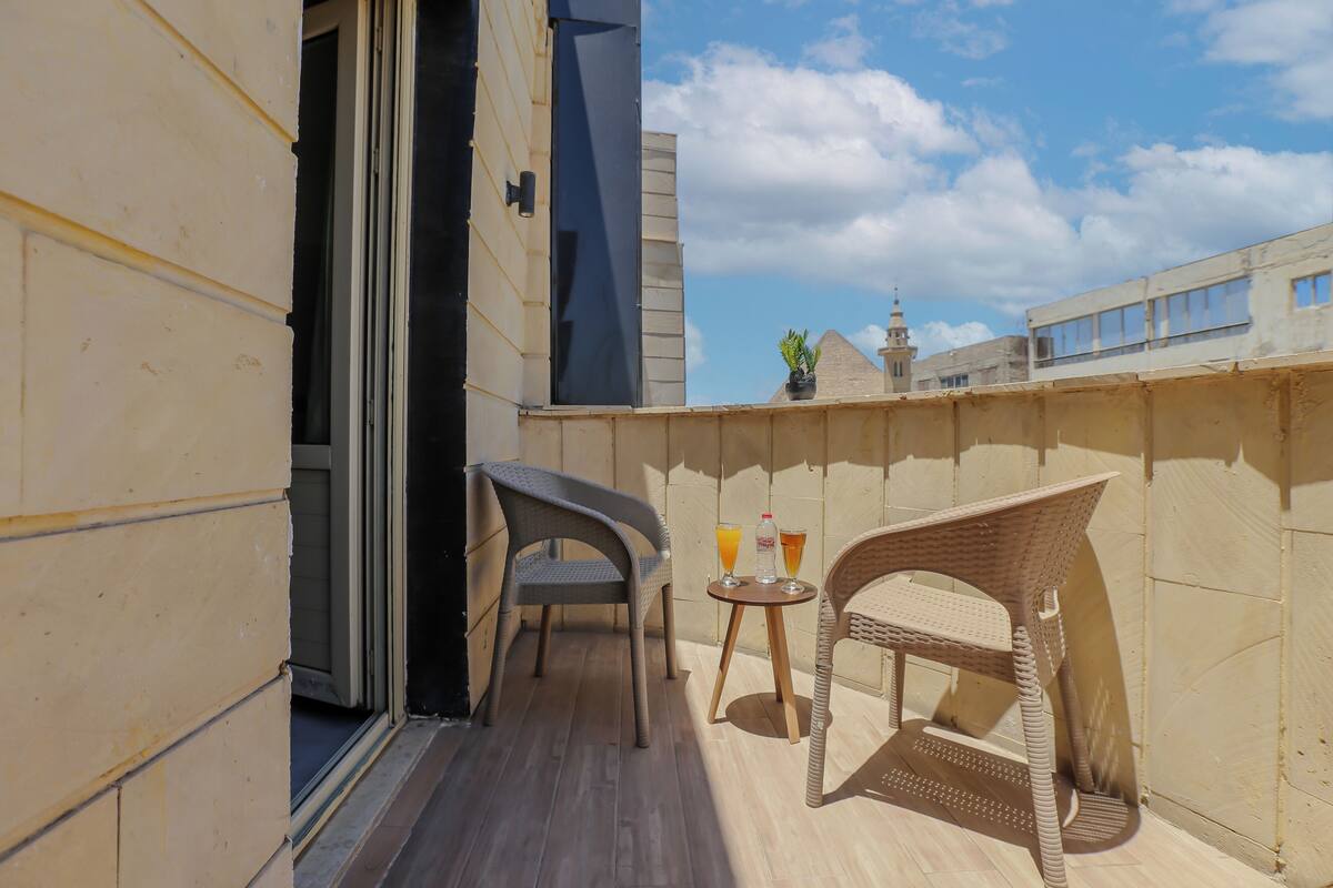A balcony with two wicker chairs, a small table with drinks, and a view of a building with a tower.