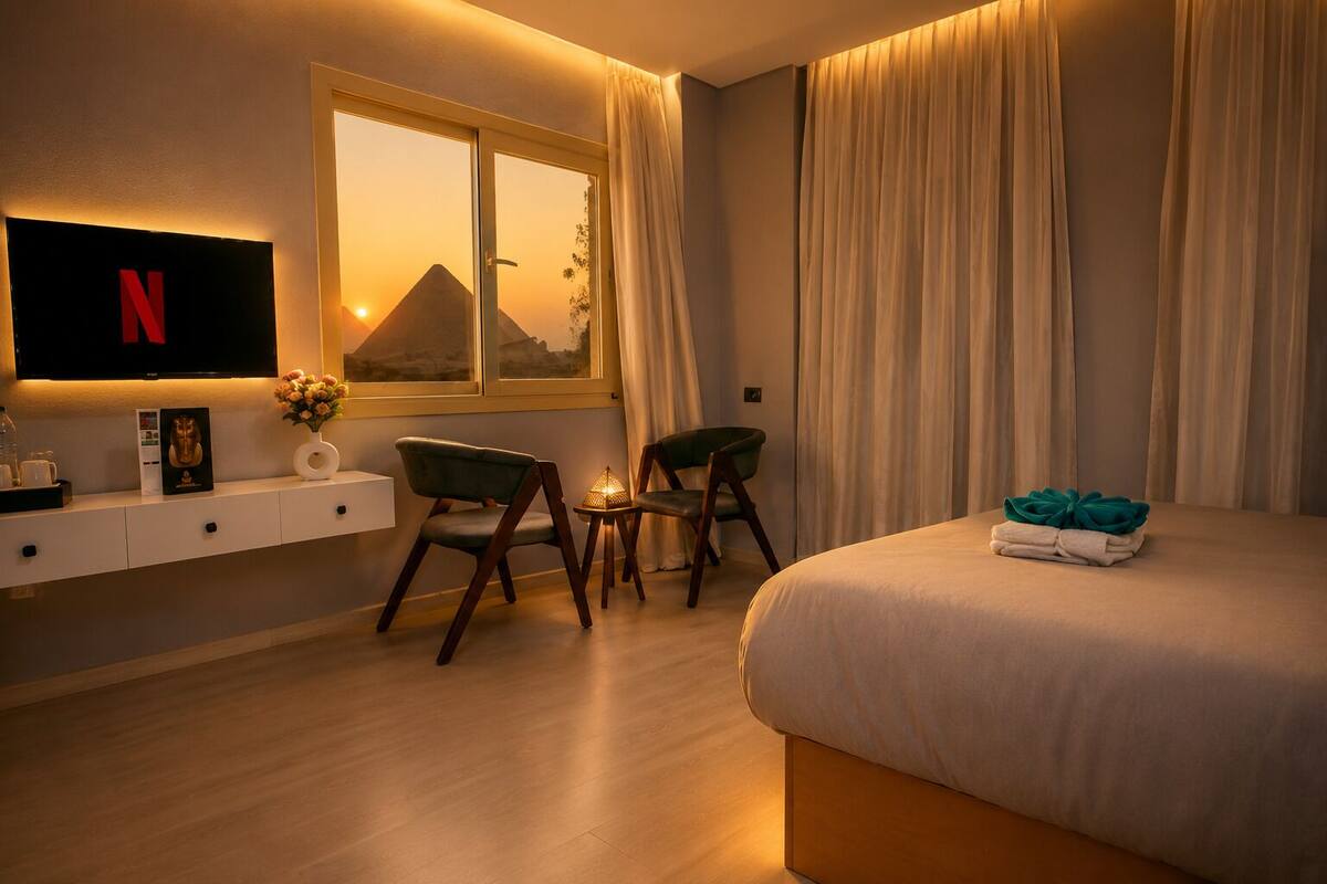 A hotel room with a bed, two chairs, a TV, a window with curtains, and a wall-mounted shelf with items.