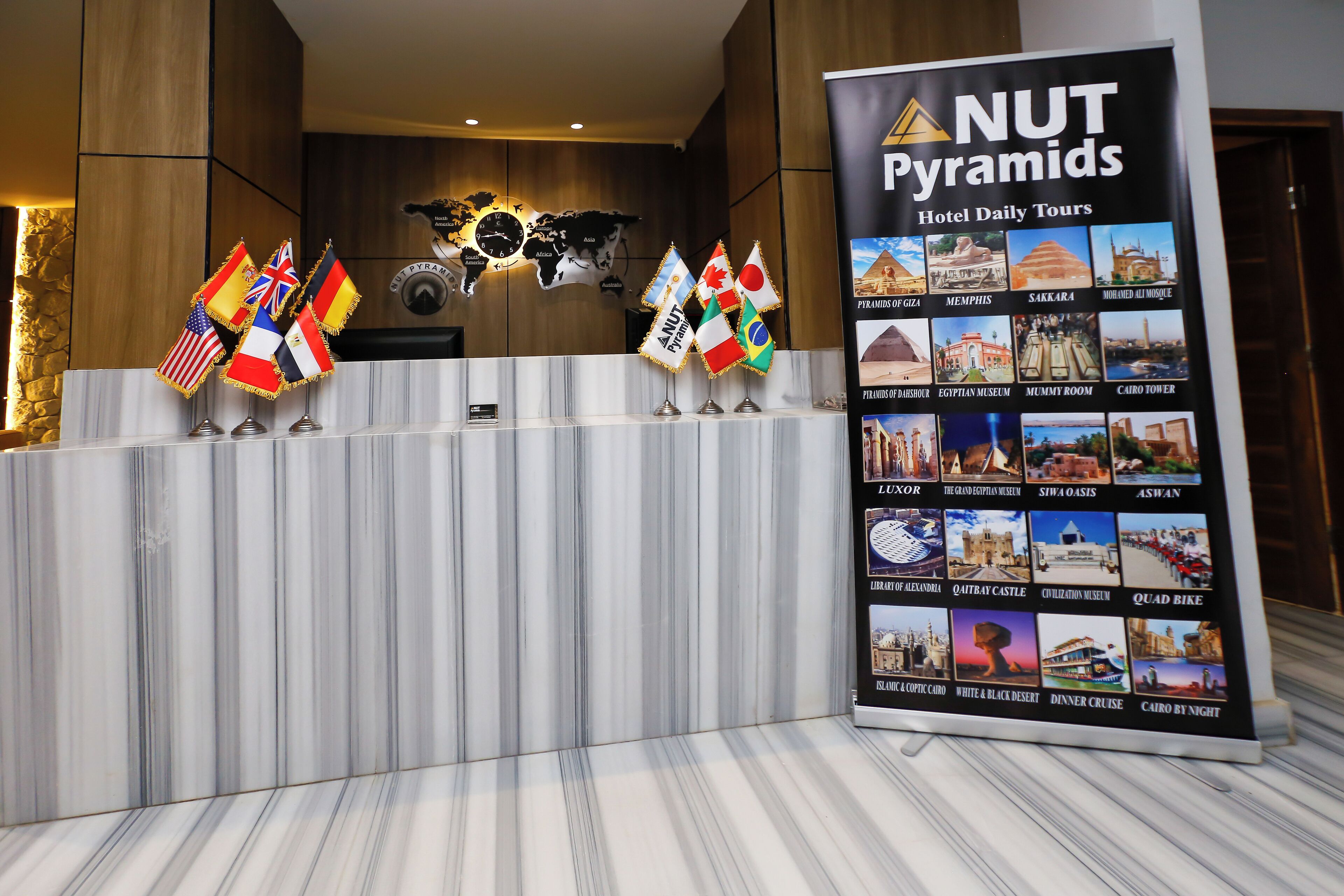 A reception desk with flags, a world map, and a sign for 'NUT Pyramids Hotel Daily Tours' listing various tour destinations.