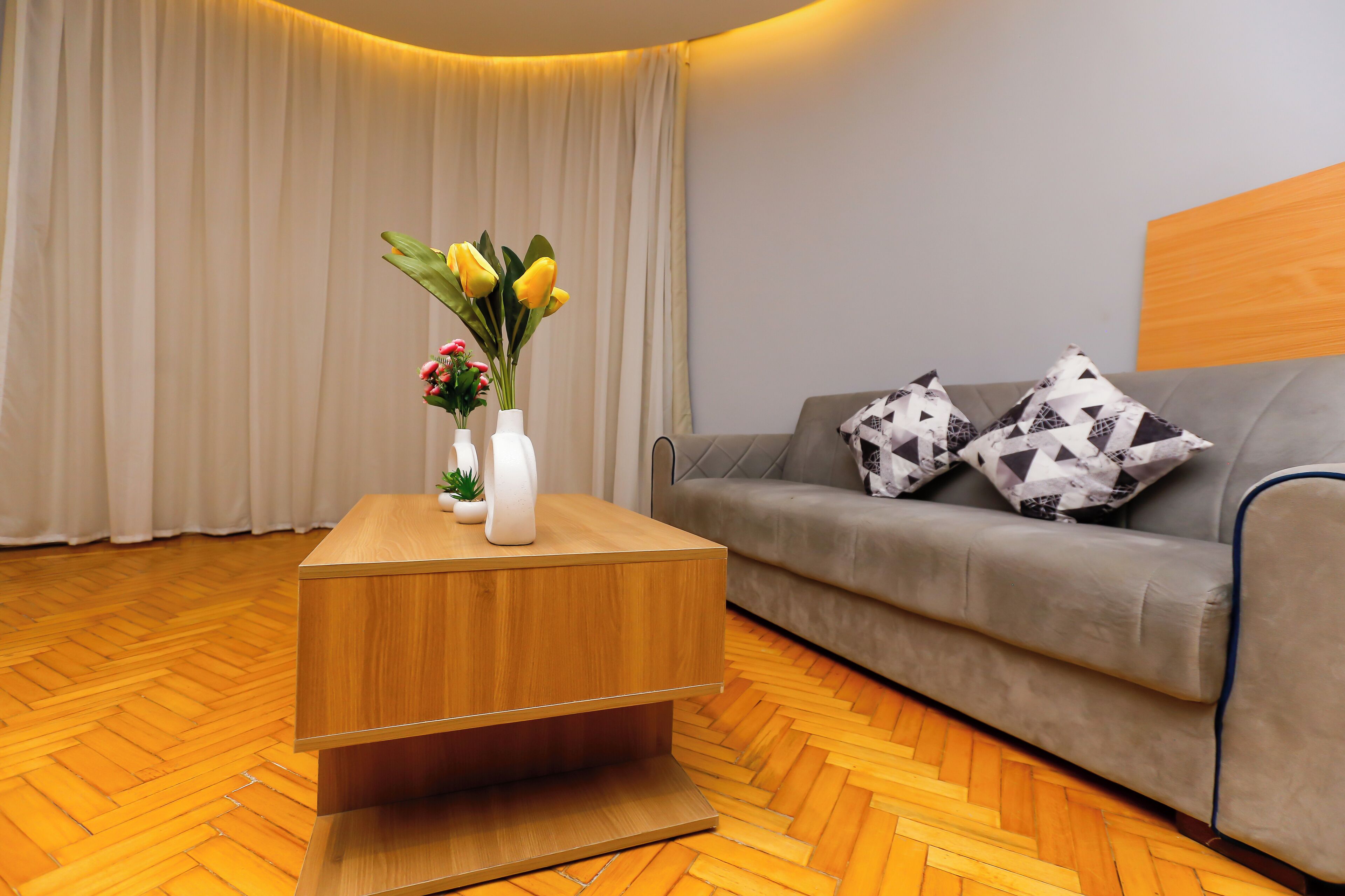 A modern living room with a wooden coffee table, a grey sofa with patterned cushions, and a vase with flowers.