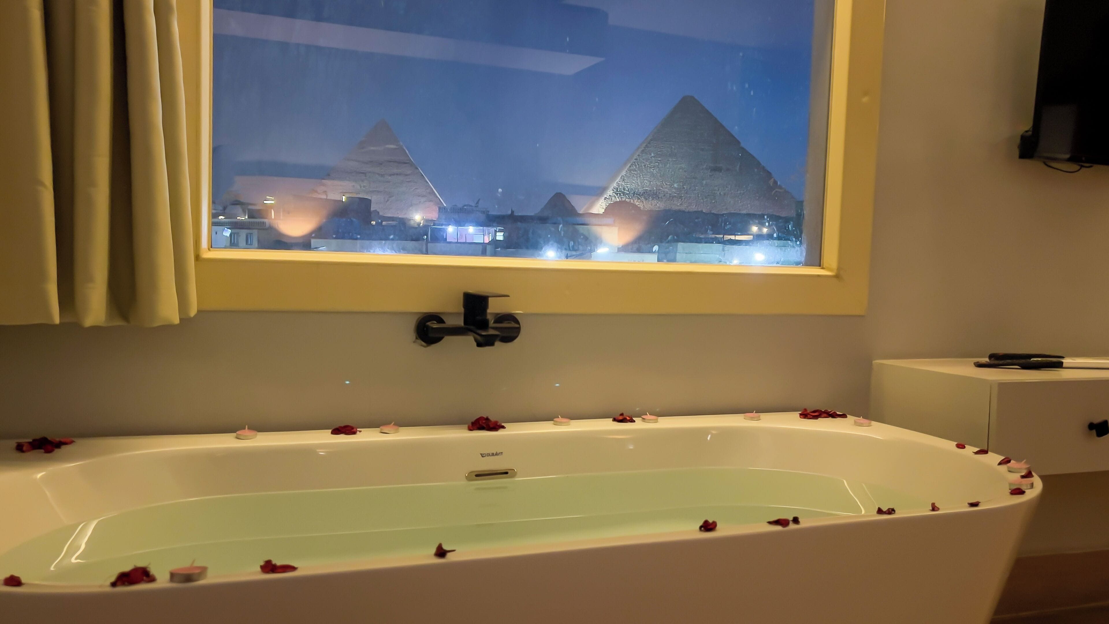 A bathtub with rose petals, a window with a view of pyramids, and a cabinet with a remote control.