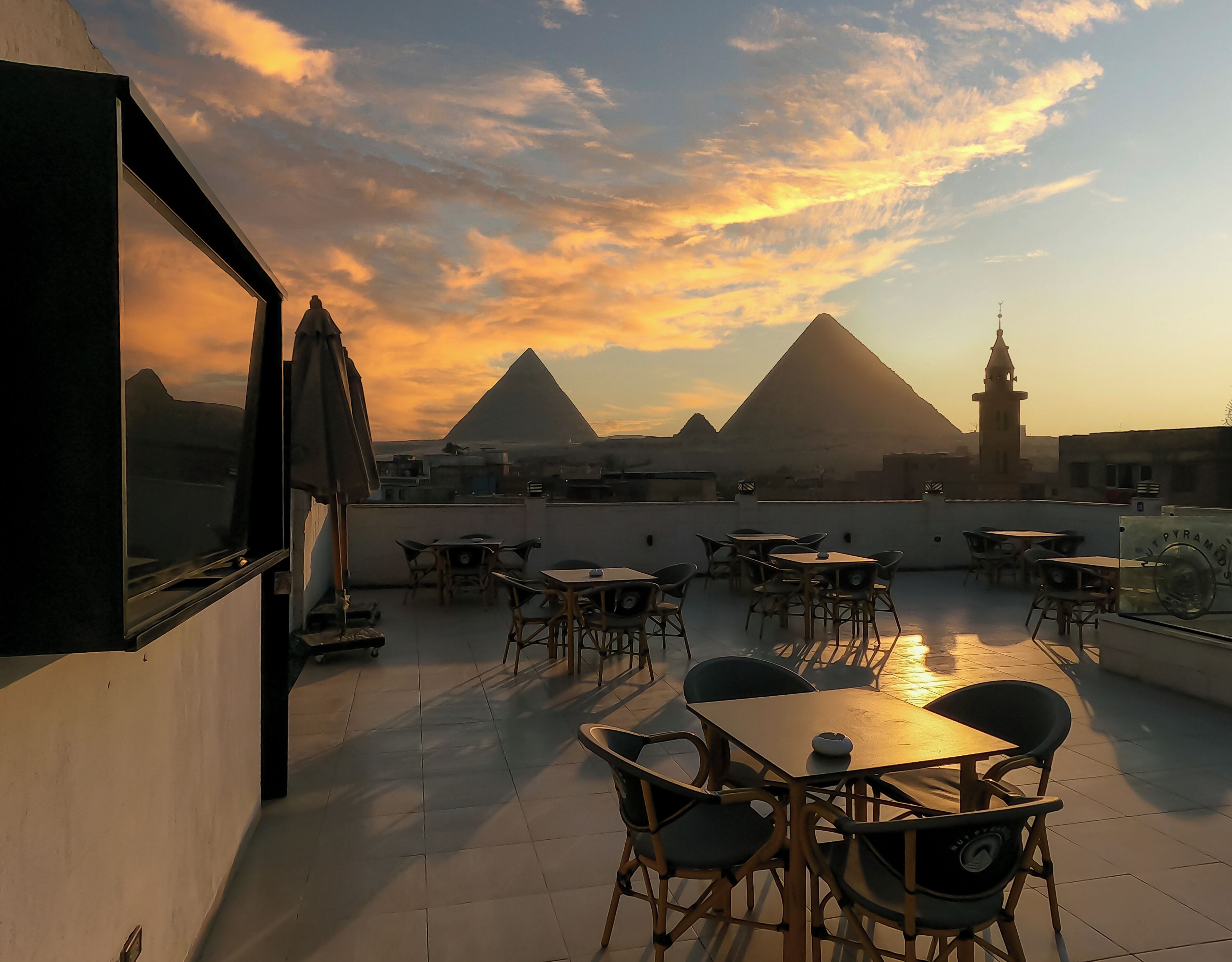 A rooftop terrace with tables and chairs, offering a view of a pyramid and a tower against a sunset sky.