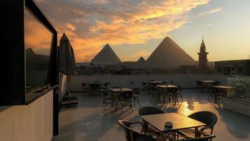 A rooftop terrace with tables and chairs, offering a view of a pyramid and a tower against a sunset sky.