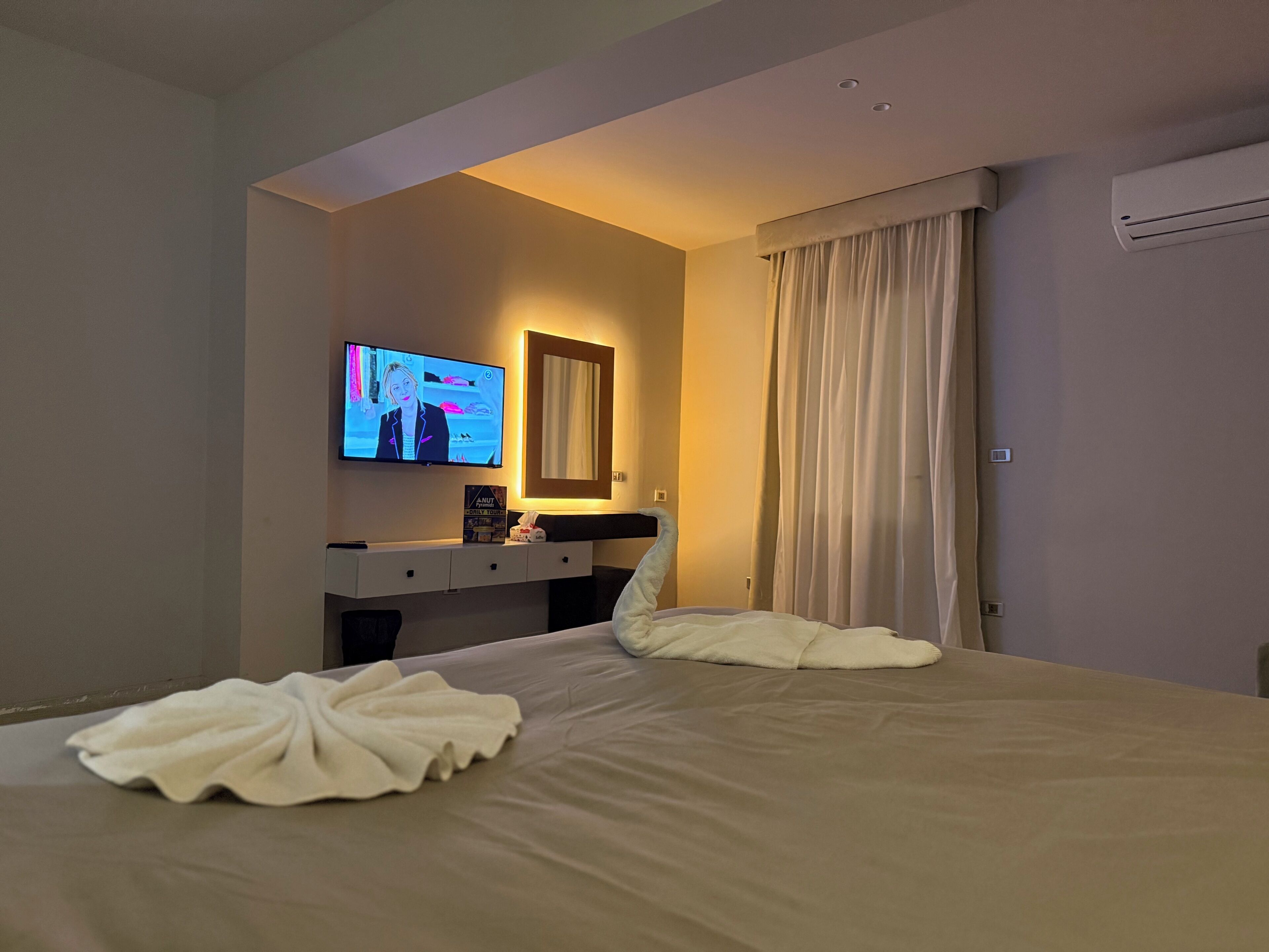 A hotel room with a bed, a TV mounted on the wall, a bedside table, and a window with curtains.