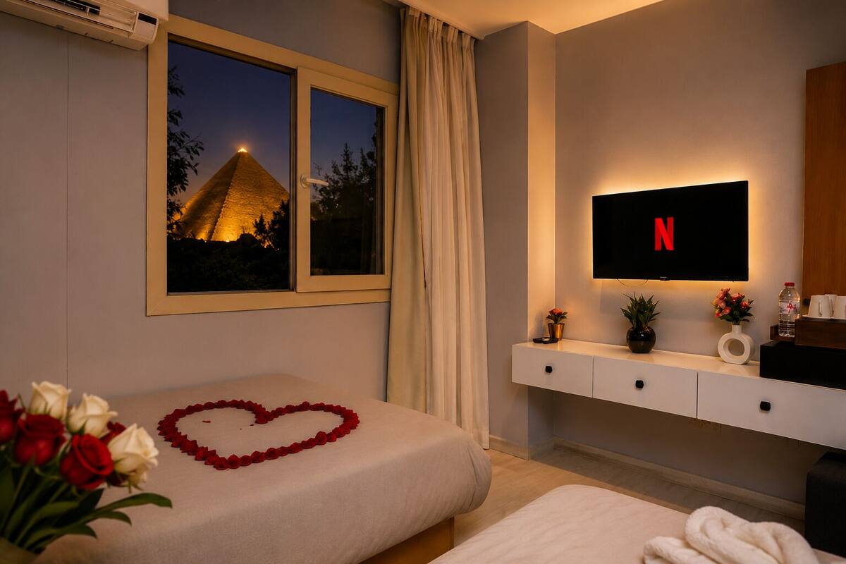 A hotel room with a bed, a TV mounted on the wall, a window with a view of a pyramid, and a bedside table with a vase of flowers.