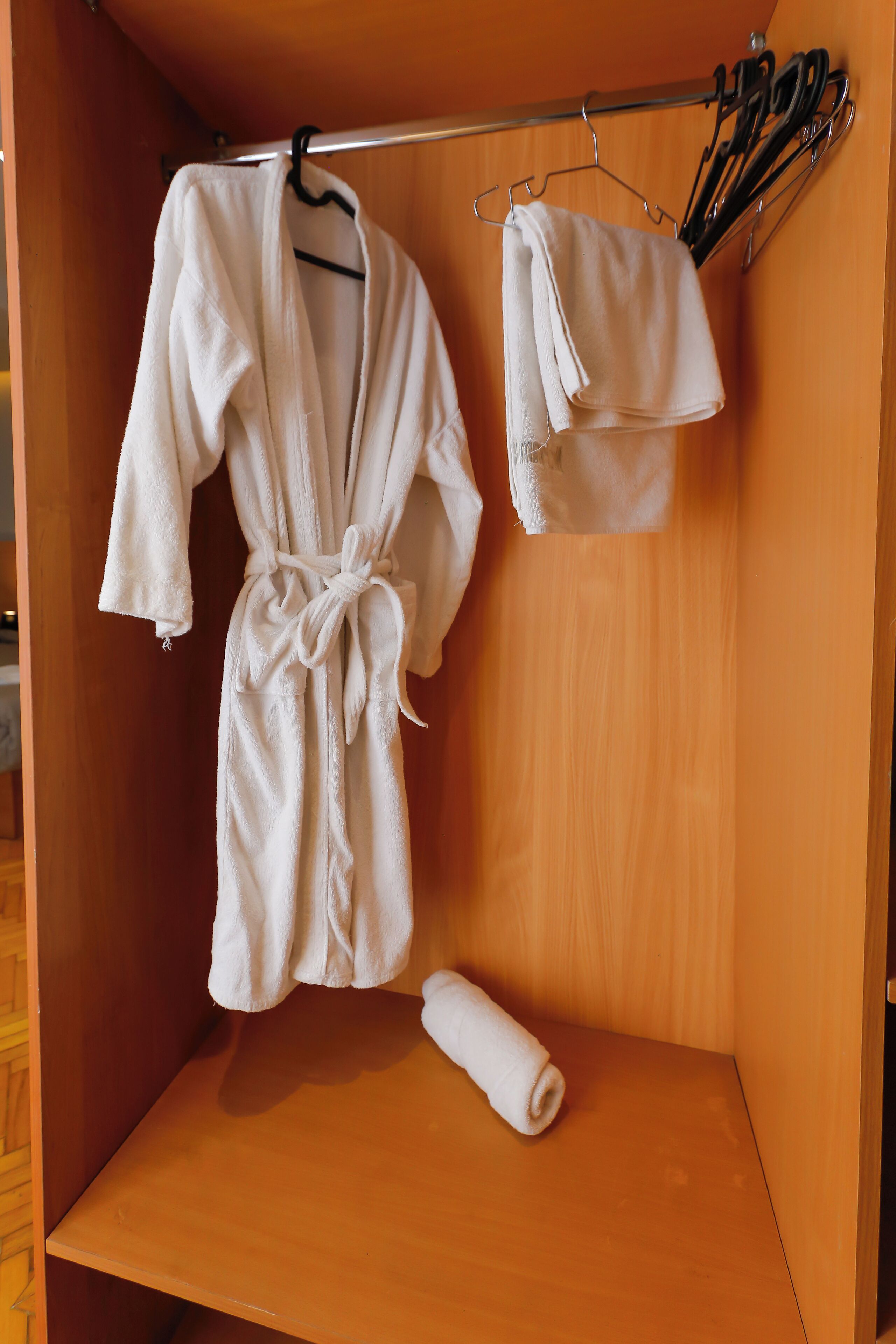 A wooden cabinet with a white robe hanging on a metal rack, a folded towel on the shelf, and a pair of scissors on the rack.
