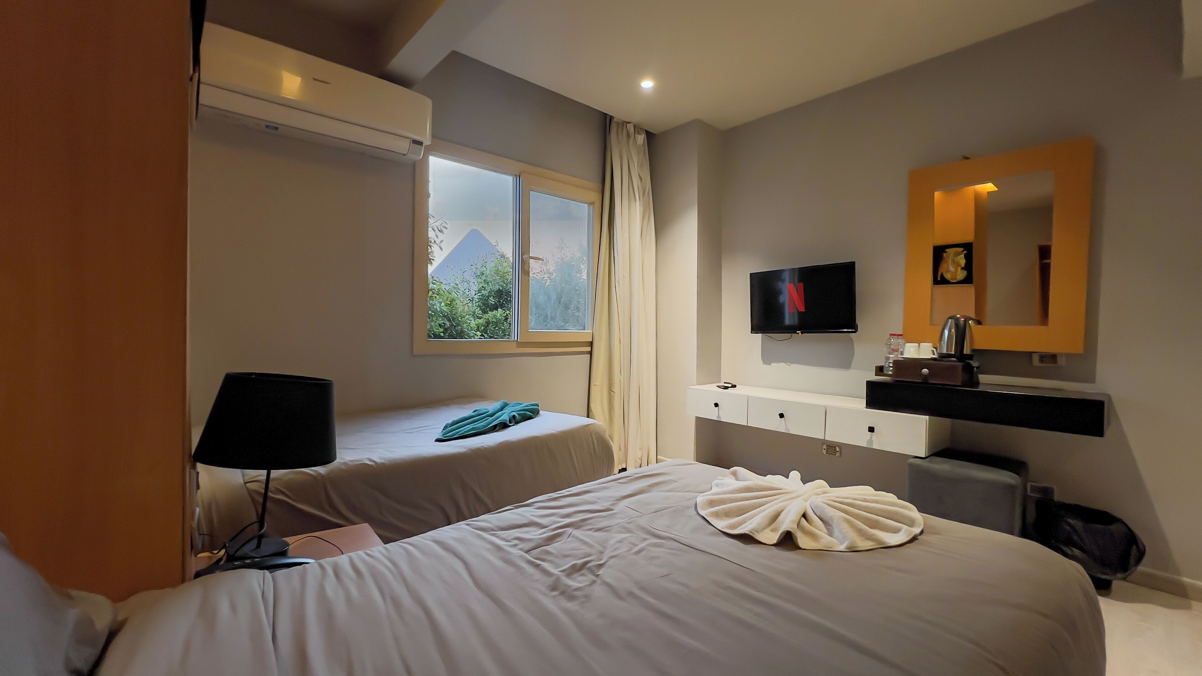 A hotel room with two beds, a TV, a mirror, and a window with curtains.