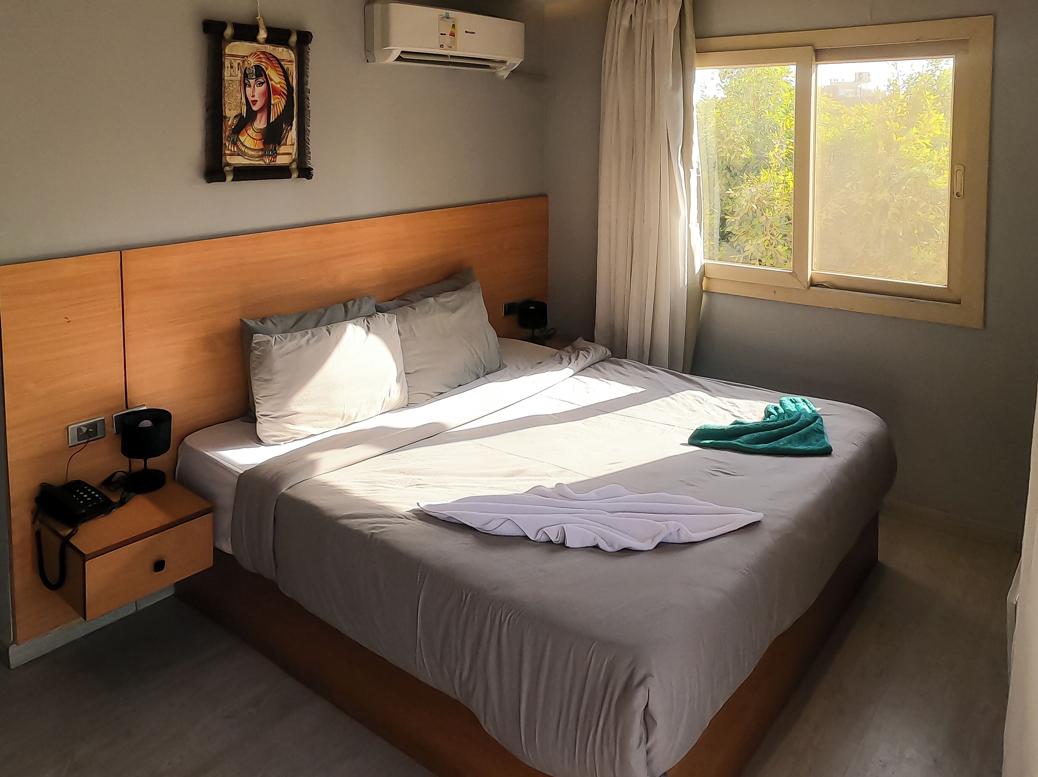Ein ordentlich bezogenes Bett mit weißer Decke und Kissen, ein Nachttisch mit Telefon und Lampe, eine Wand-Klimaanlage und ein Fenster mit Vorhängen.