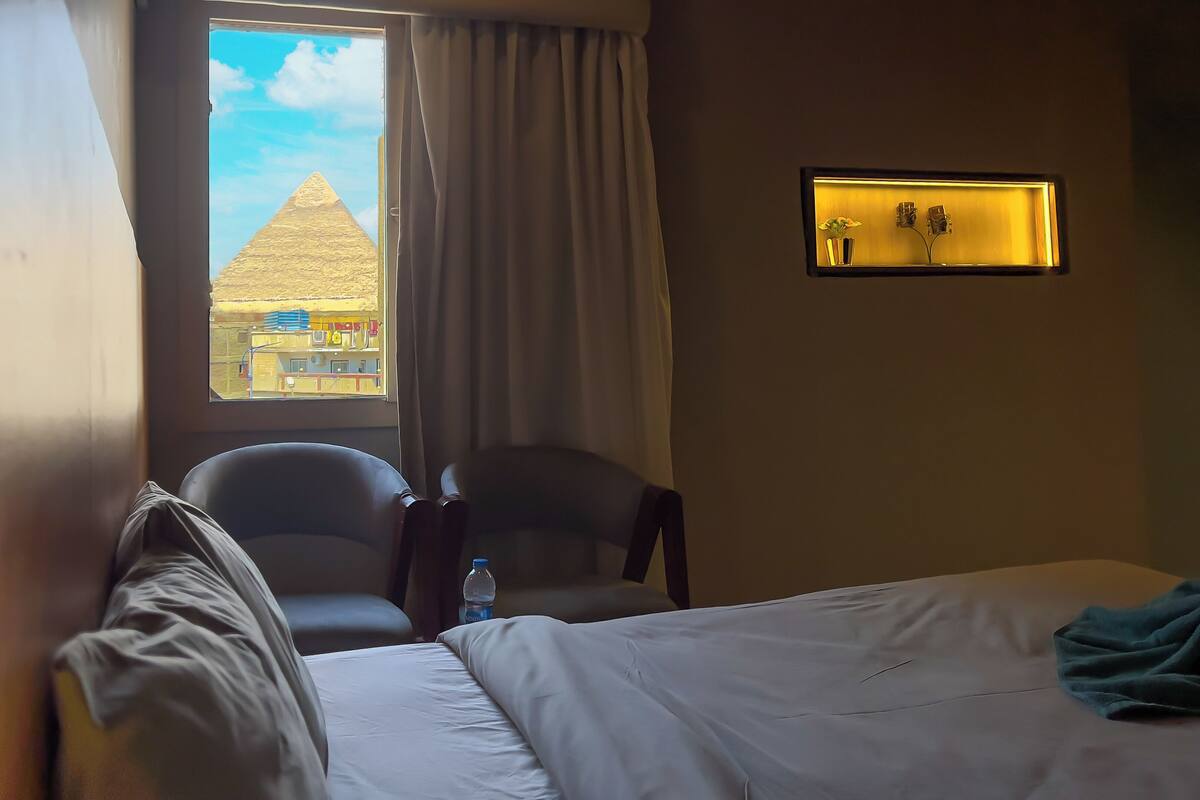 A hotel room with a bed, a chair, a window with a view of a pyramid, and a wall-mounted light fixture.