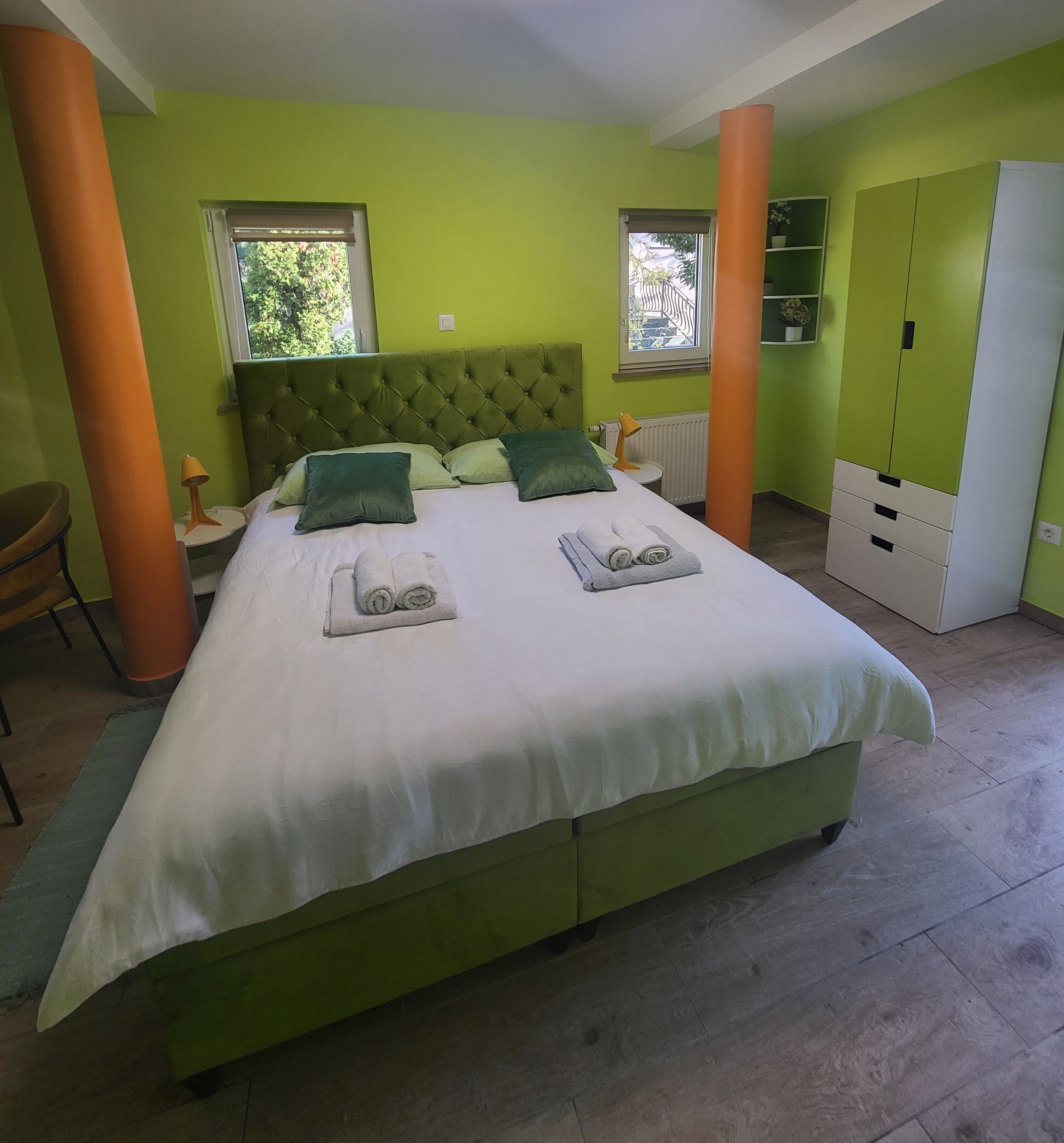 A room with a green upholstered bed, two pillows, a white duvet, and two rolled towels on the bed. There are two windows with blinds, a white radiator, and a green wardrobe with a white door.