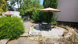 Een terras met een tafel en stoelen, omgeven door groen en een gebouw.