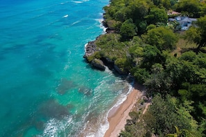 Vue aérienne d’une zone côtière avec une plage, une végétation luxuriante et une maison dotée d’un toit en pente.