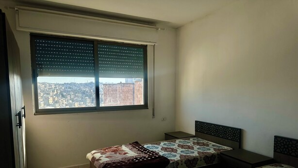 A bedroom with two beds, a window with blinds, and a city view.