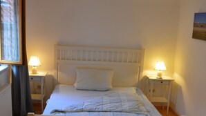 Une chambre avec un lit simple, une tête de lit blanche, deux tables de chevet avec des lampes, une fenêtre avec des rideaux et un radiateur.