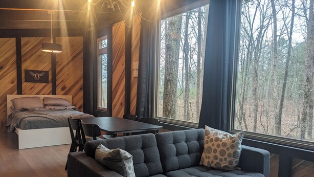 A cozy cabin interior with a bed, sofa, dining table, and large windows offering a forest view.