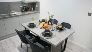 A modern kitchen with grey tiles, a stainless steel sink, and a dining table set with plates, glasses, and a fruit platter. - City Centre Luxury ApartmentSofa BedWIFI (Leicester)