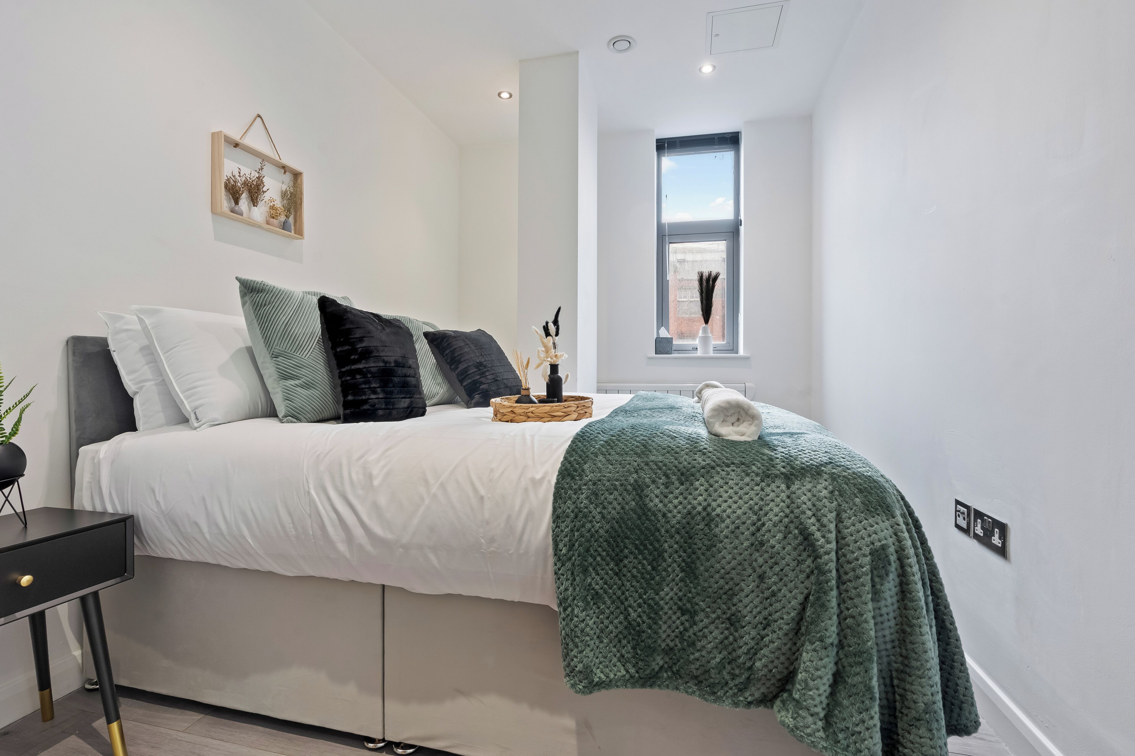 A modern bedroom with a bed, pillows, a bedside table, a window, and a wall-mounted light fixture.