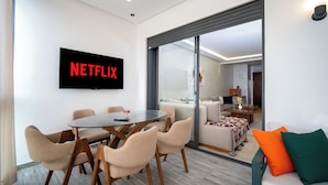 Een moderne woonkamer met een glazen scheidingswand, een ronde eettafel met stoelen, een bank met kussens en een flatscreen televisie waarop het Netflix logo staat.