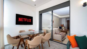 Un salon moderne avec une cloison en verre, une table de salle à manger ronde entourée de chaises, un canapé avec des coussins et un téléviseur à écran plat affichant le logo de Netflix.