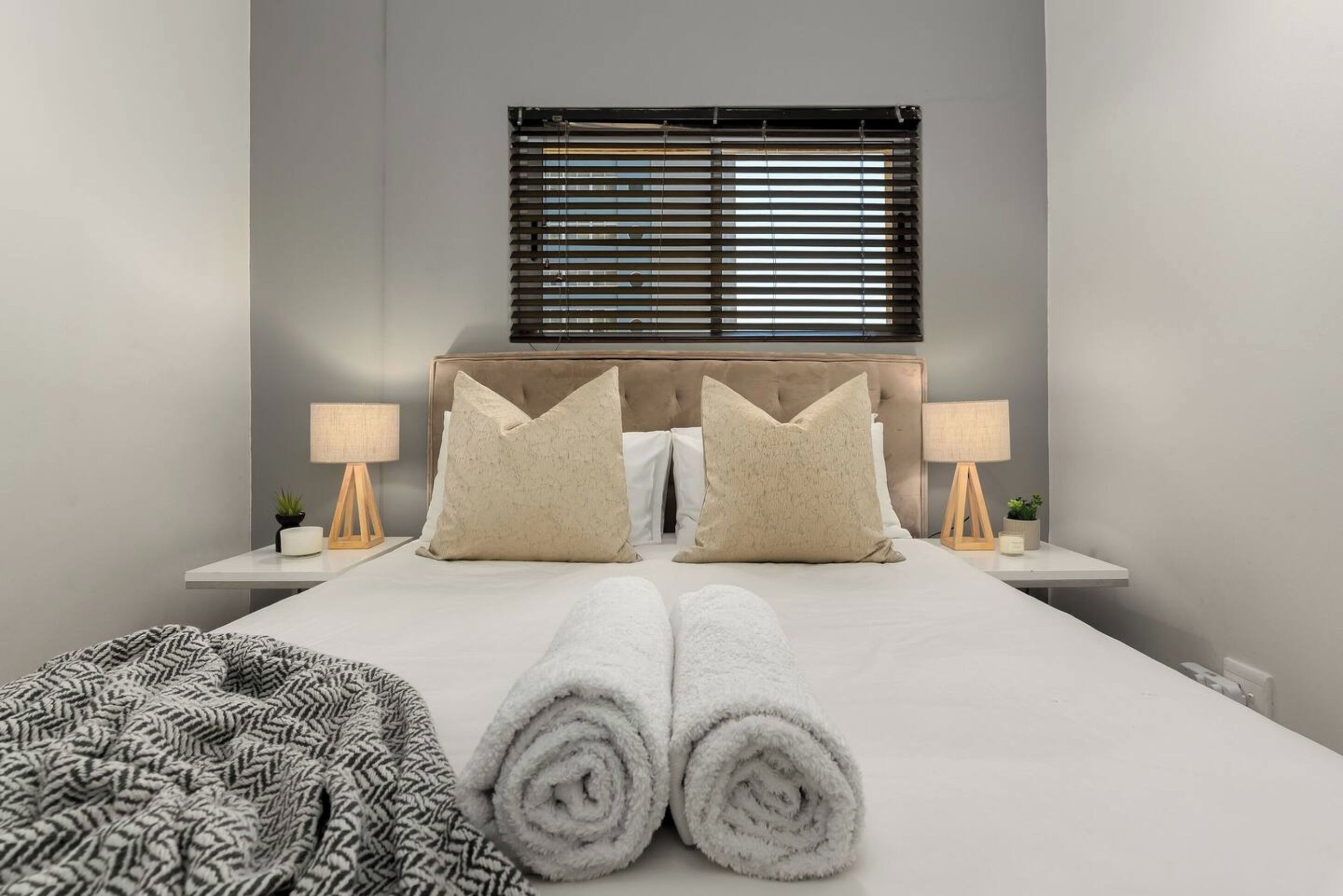 Una cama bien hecha con toallas enrolladas, dos mesitas de noche con lámparas y una ventana con persianas.