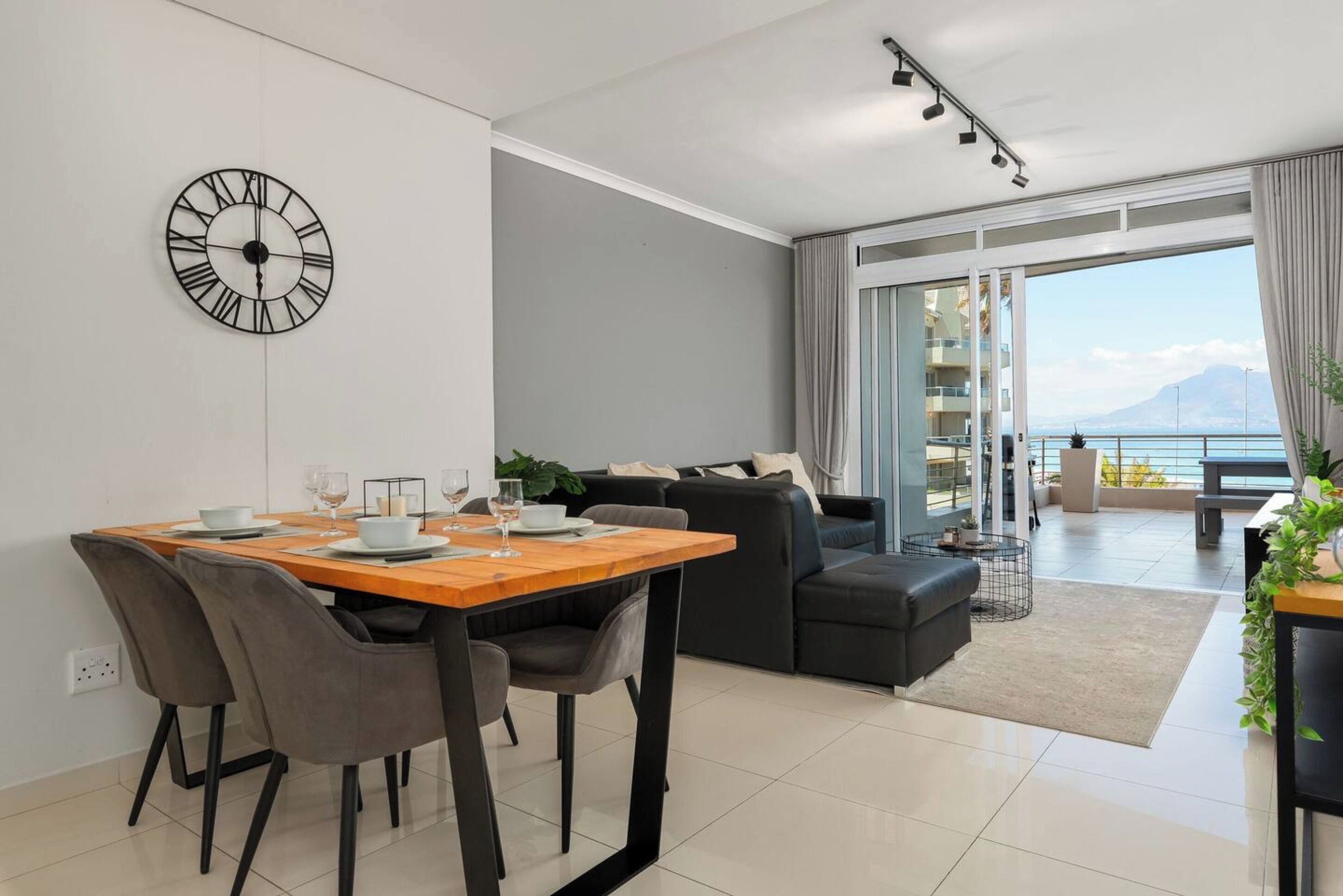 Ein modernes Wohnzimmer mit Essbereich, einem großen Fenster mit Meerblick und einer schwarzen Wanduhr.