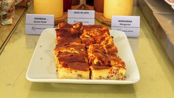 Una presentación de postres con un plato de rebanadas, dos frascos etiquetados como 'GOIABADA' y 'MARGARINA', y una cuchara de madera.