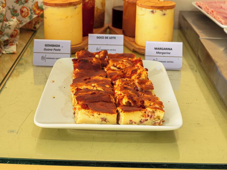 Una presentación de postres con un plato de rebanadas, dos frascos etiquetados como 'GOIABADA' y 'MARGARINA', y una cuchara de madera.