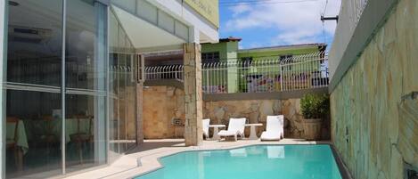 A swimming pool with lounge chairs, a stone wall, and a building with large windows and a sign.