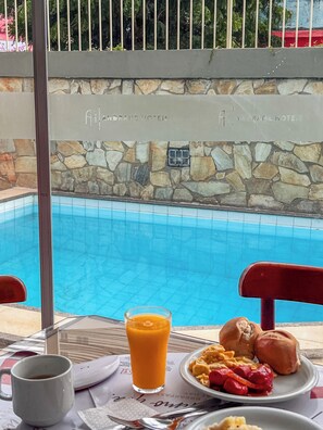 A dining table with breakfast items and a glass of orange juice, overlooking a pool with a stone wall and a sign reading "Andrade Hotels". - Hotel Jangadeiro (Aracaju)