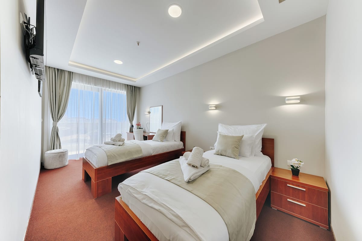 A hotel room with two beds, a bedside table with a lamp, a large window with curtains, and a wall-mounted air conditioning unit.