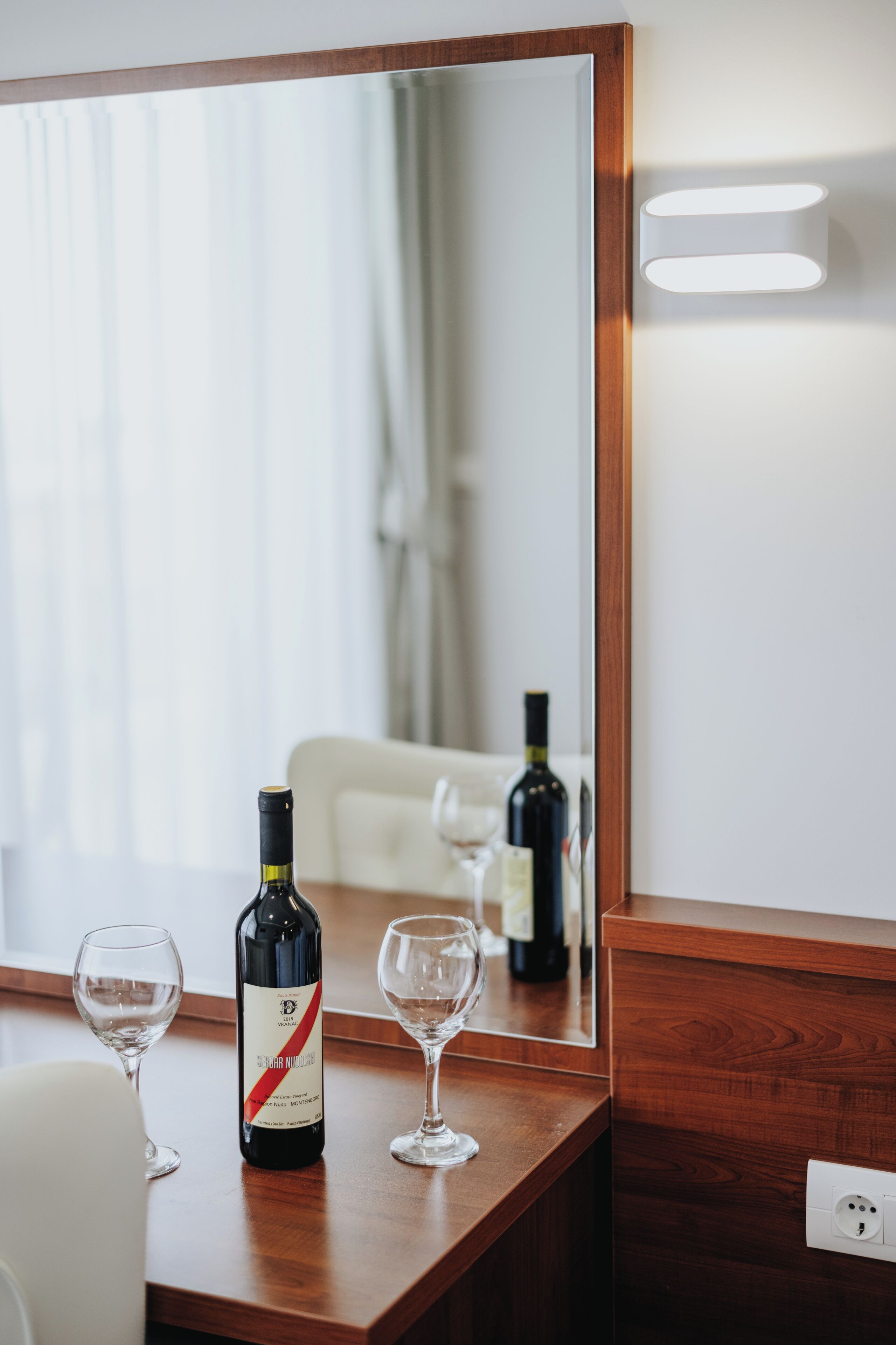 A wooden table with a wine bottle and two glasses, a mirror reflecting a room with a white chair and curtains.