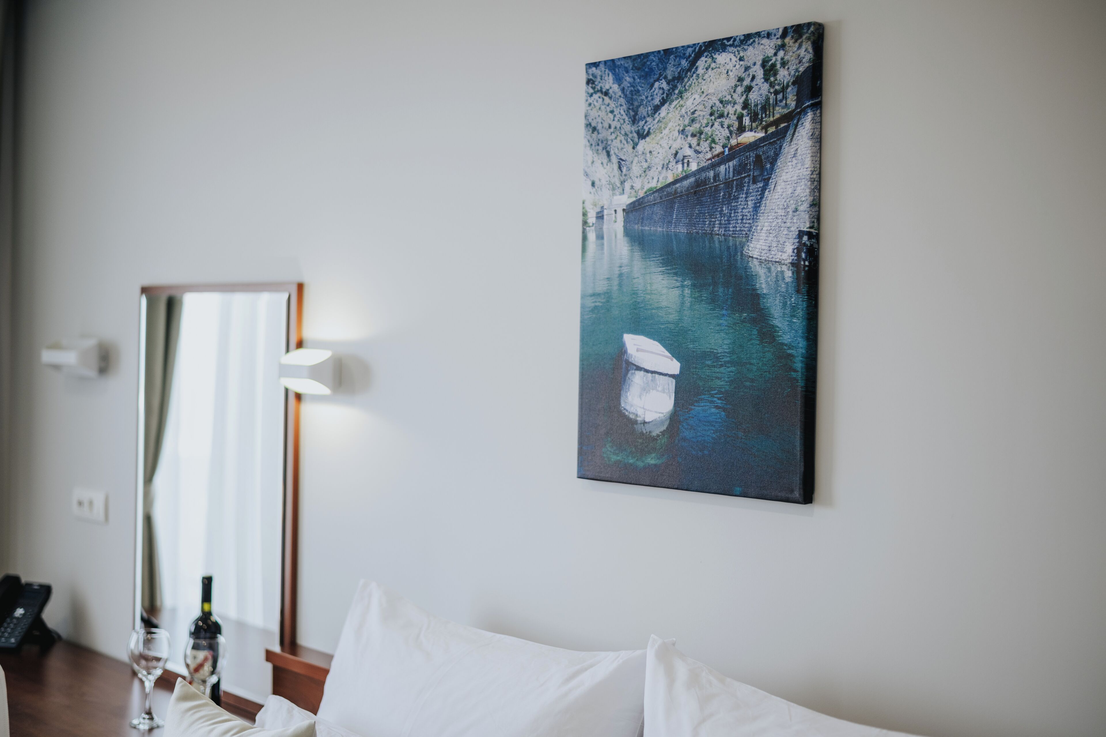 A hotel room with a bed, a bedside table with a bottle and glass, a painting of a bridge over water, and a mirror on the wall.