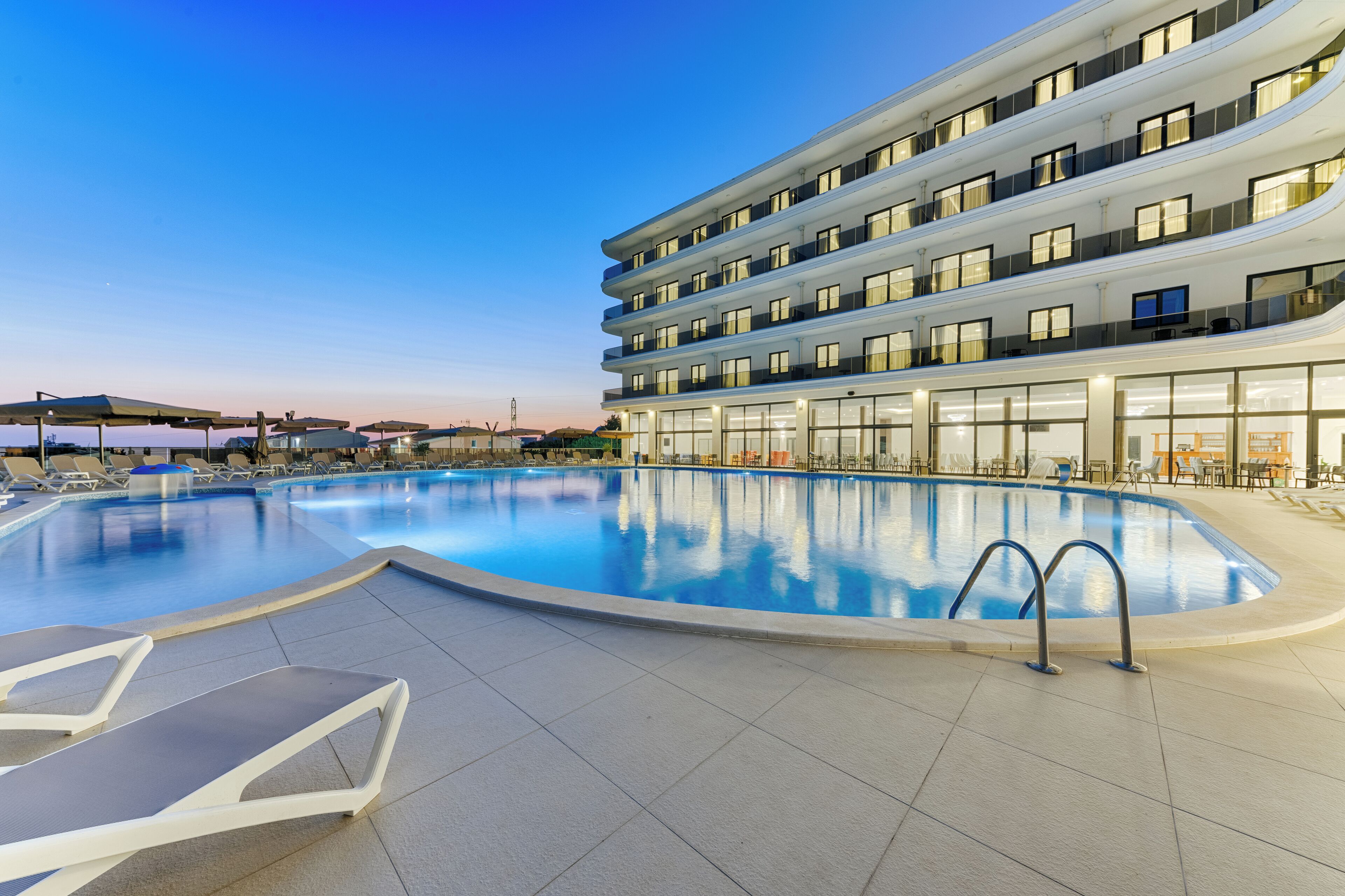 A modern hotel with a large outdoor swimming pool, multiple balconies, and a well-lit facade.