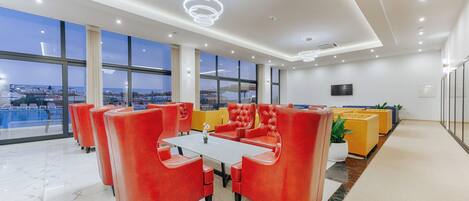 A modern hotel lobby with a large seating area, a conference table, and a view of the cityscape through large windows.
