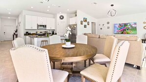 Una sala de estar moderna con una mesa de comedor redonda de madera, rodeada por cuatro sillas tapizadas. Se ve un sofá y un televisor al fondo.