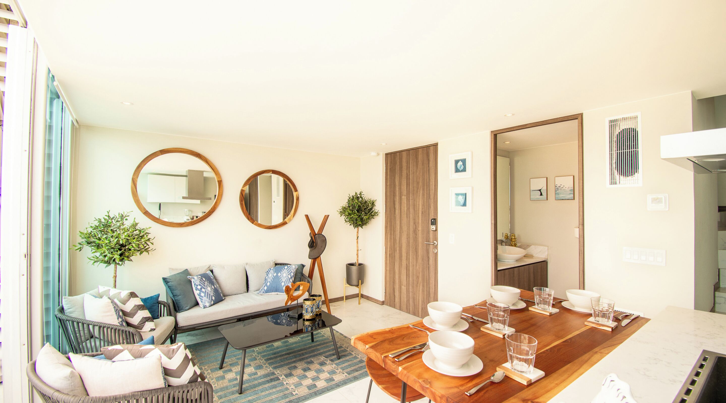 A modern living room with a sofa, coffee table, and dining table set for four. There are two mirrors on the wall, a potted plant, and a door leading to a bathroom.