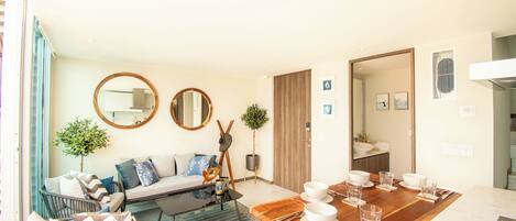A modern living room with a sofa, coffee table, and dining table set for four. There are two mirrors on the wall, a potted plant, and a door leading to a bathroom.