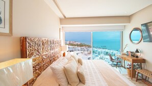 Un dormitorio con una cama grande, un escritorio con silla, vista al mar y al paisaje urbano.