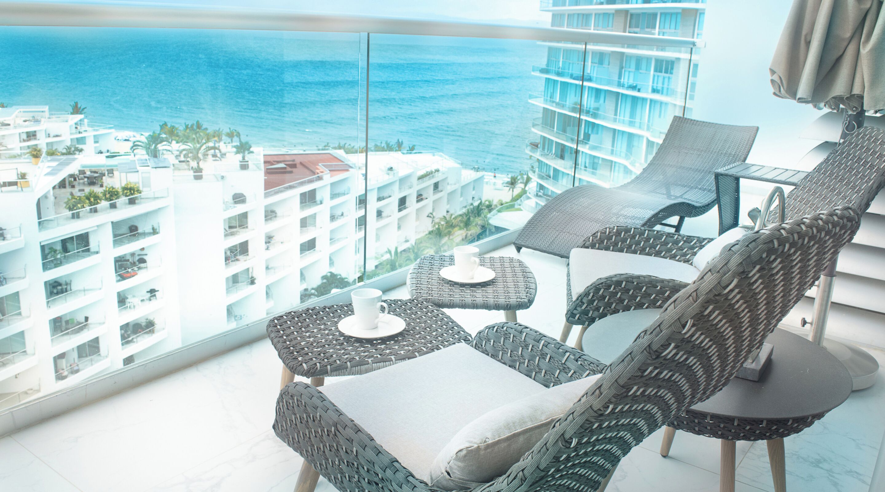 Een balkon met rieten meubilair, een glazen tafel en uitzicht op zee en gebouwen.