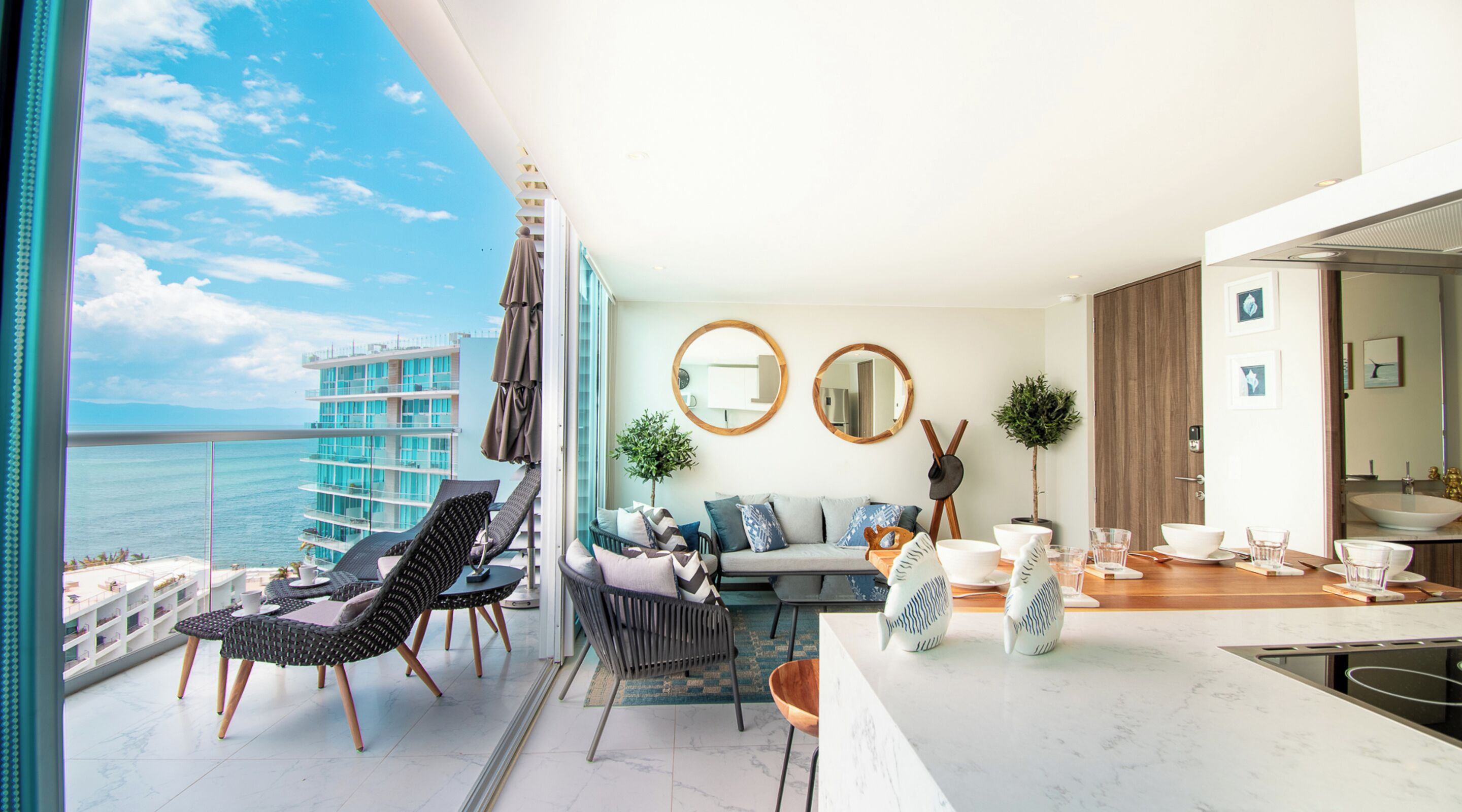 Een moderne woonkamer met balkon en uitzicht op zee, een eethoek met keuken, en een zicht op een hoog gebouw.