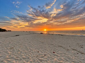 Een strand met een ondergaande zon, zand en een verre zeilboot.