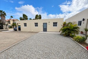 Un edificio de dos pisos con camino de grava, palmeras y cielo azul despejado.