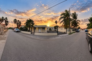 A street view at sunset with a gated building, palm trees, and parked cars. - Aruba IXORA H2 - Swalchi Suite, studio, sleeps 2, 7 minutes drive to the beach (Noord)