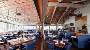 Restaurante com pé-direito alto, vigas aparentes e um espaço de bar. Mesas organizadas com cadeiras azuis.