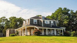 Uma casa de dois andares com varanda, cercada por um gramado bem cuidado e árvores.