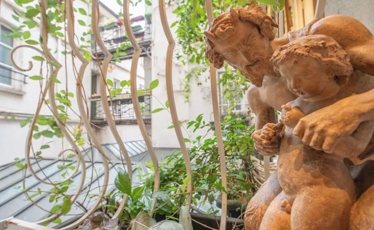 Una escultura de terracota con querubines en un balcón, con plantas y un edificio al fondo.
