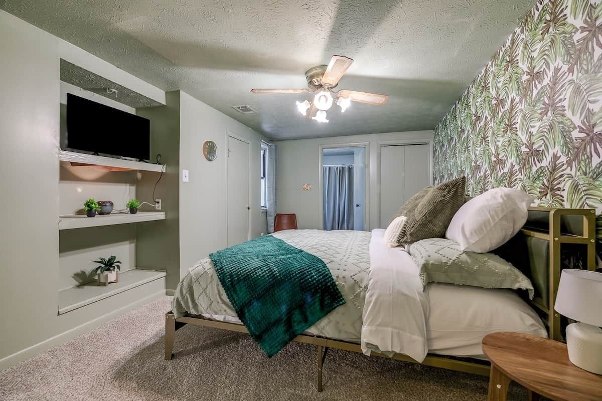 A bedroom with a bed, a ceiling fan, a TV mounted on the wall, and a window with curtains.