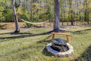 A hammock between trees, a fire pit, and a wooden bench in a forest setting.