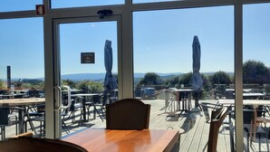 Zona d'esmorzar a l'aire lliure amb taules i caires, vista del paisatge i un senyal que indica un restaurant.